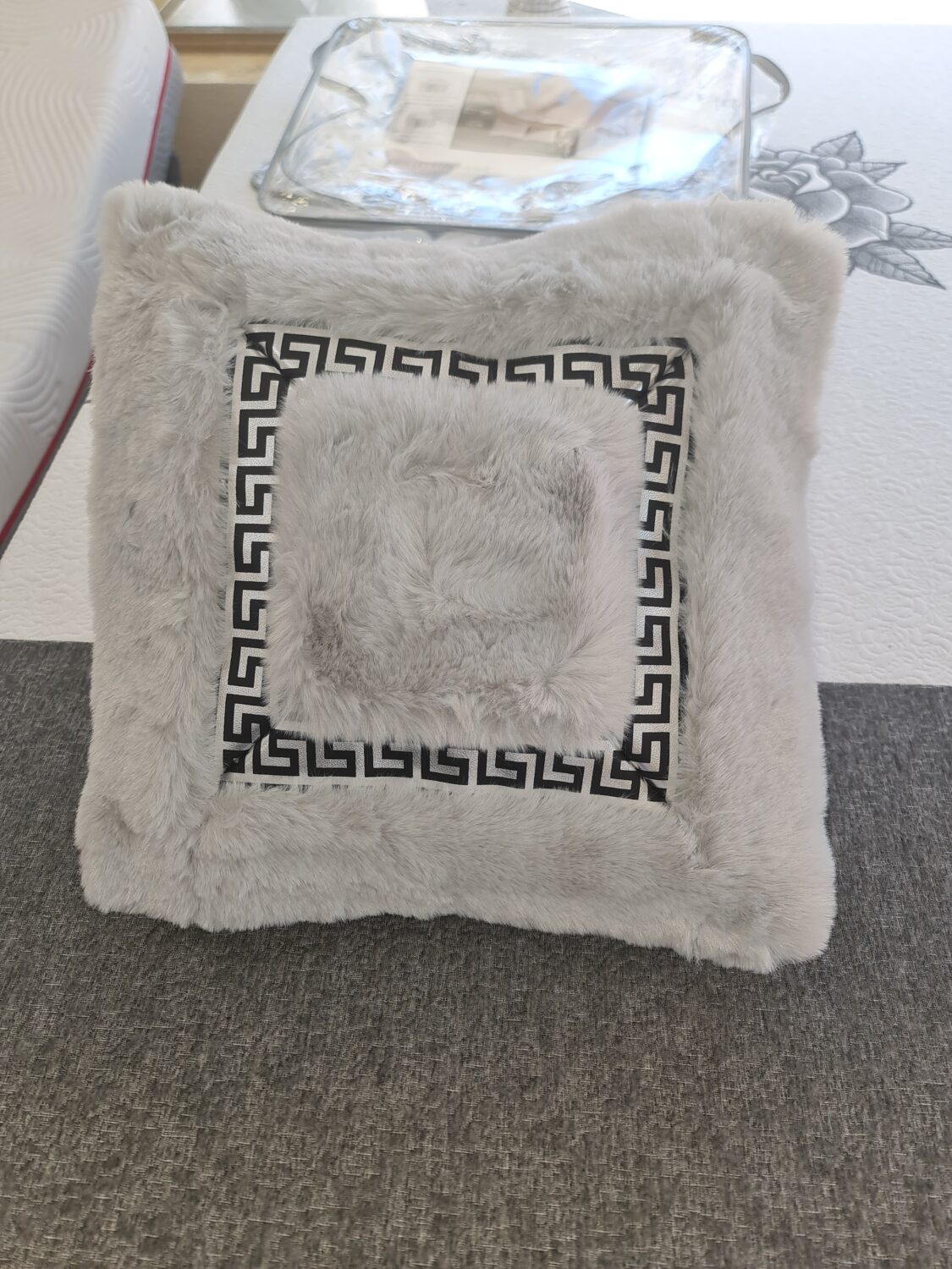 Cojín decorativo de piel sintética gris presenta un diseño de greca griega negra en el centro y una textura suave y esponjosa